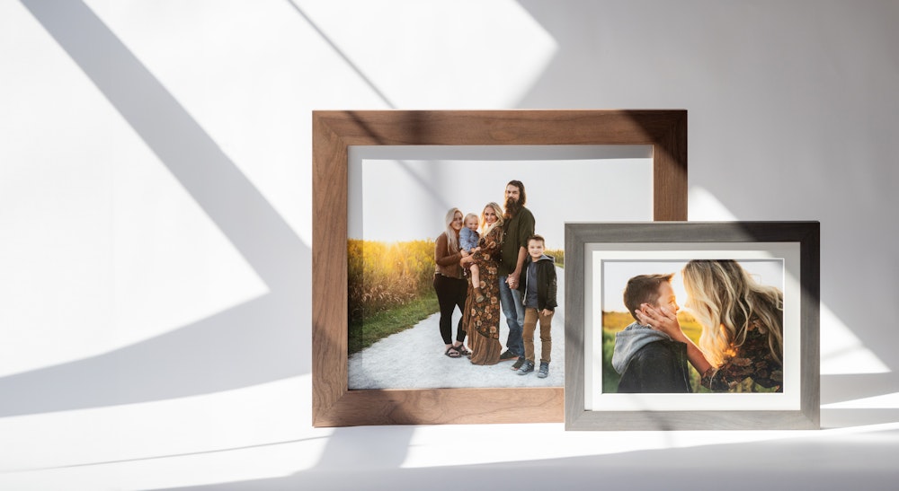 Two picture frames on a white surface with shadows from window light. The large walnut Woodland frame has a family photo with five people standing outdoors. The smaller gray Woodland frame shows a close-up of an adult kissing a child's cheek in a sunny setting.