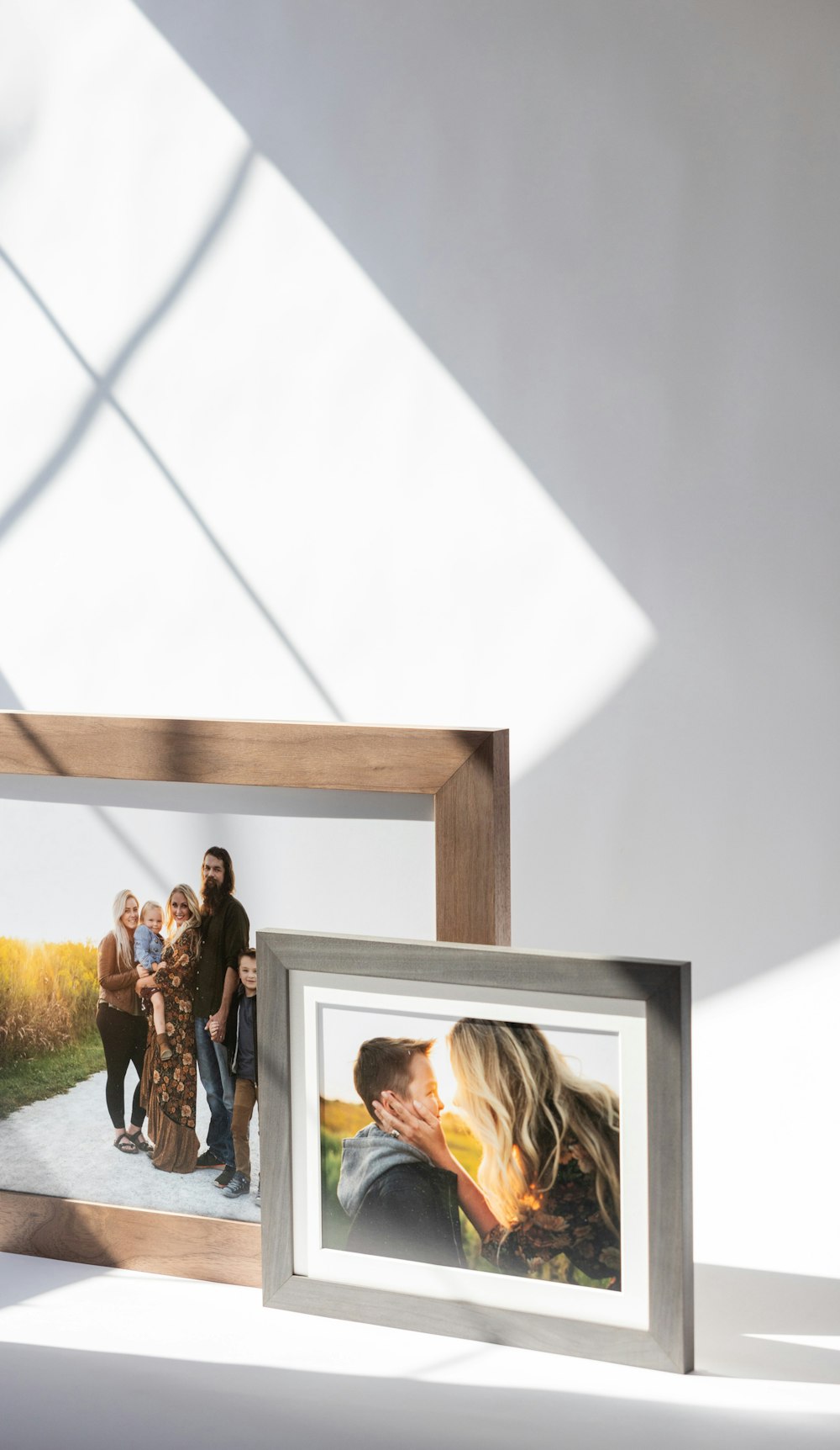 Two picture frames on a white surface with shadows from window light. The large walnut Woodland frame has a family photo with five people standing outdoors. The smaller gray Woodland frame shows a close-up of an adult kissing a child's cheek in a sunny setting.