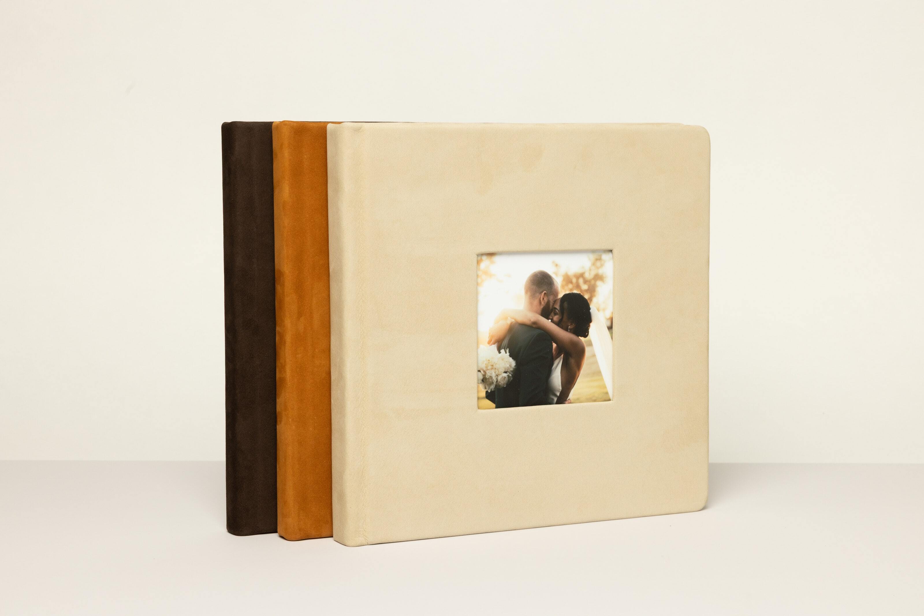 Three 10×10 albums are standing upright in a row featuring the new Nubuck Leathers with the front Album showing a wedding couple in a Ecru Nubuck Leather Cameo Cover.