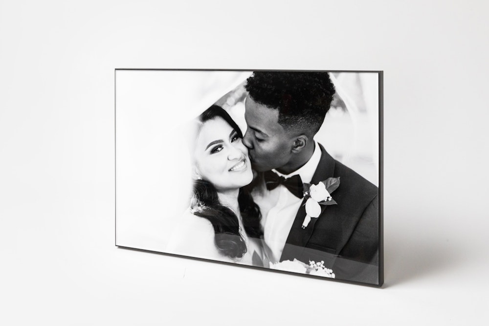 Wedding portrait of groom kissing bride's cheek as veil floats around them, displayed in a Black Framed Acrylic Print standing on a soft gray background