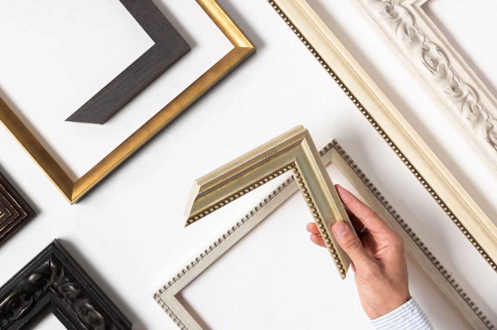 A hand holds a gold picture frame corner sample among various frame styles on a white background.