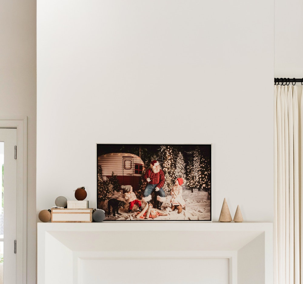 Float Framed print above a white fireplace mantel with small decorative objects, framed photograph shows a festive winter scene with people, a camper, and dogs.