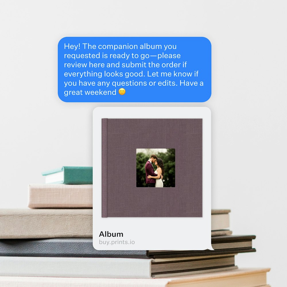 A text message notification about a ready photo album on a stack of albums and books: the message asks to review and submit the order. The album cover shows a photo of a couple hugging on their wedding day.