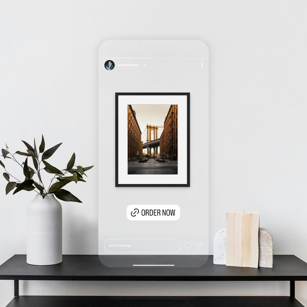 A framed photo of an urban street scene is displayed on a minimalistic shelf with a plant and books. A digital overlay with "ORDER NOW" button is visible on the photo to appear like and Instagrm Story shopping link.