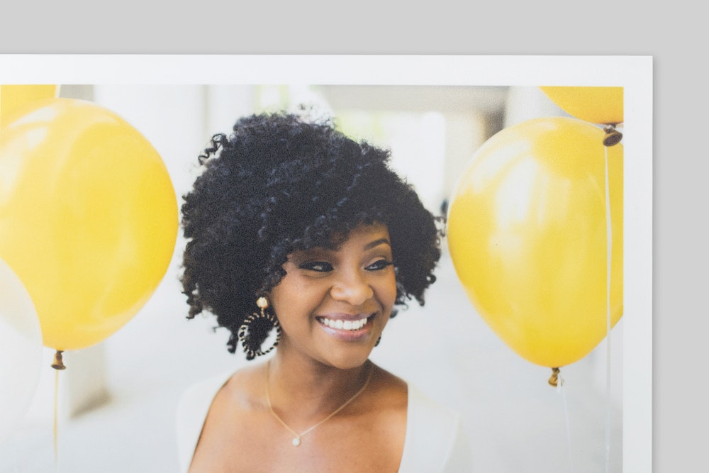 Corner of Lustre with Protective Lamination Photo Print with border, featuring a portrait of a smiling woman with curly hair amid bright yellow balloons. Subtle texture with satin finish visible on paper surface.