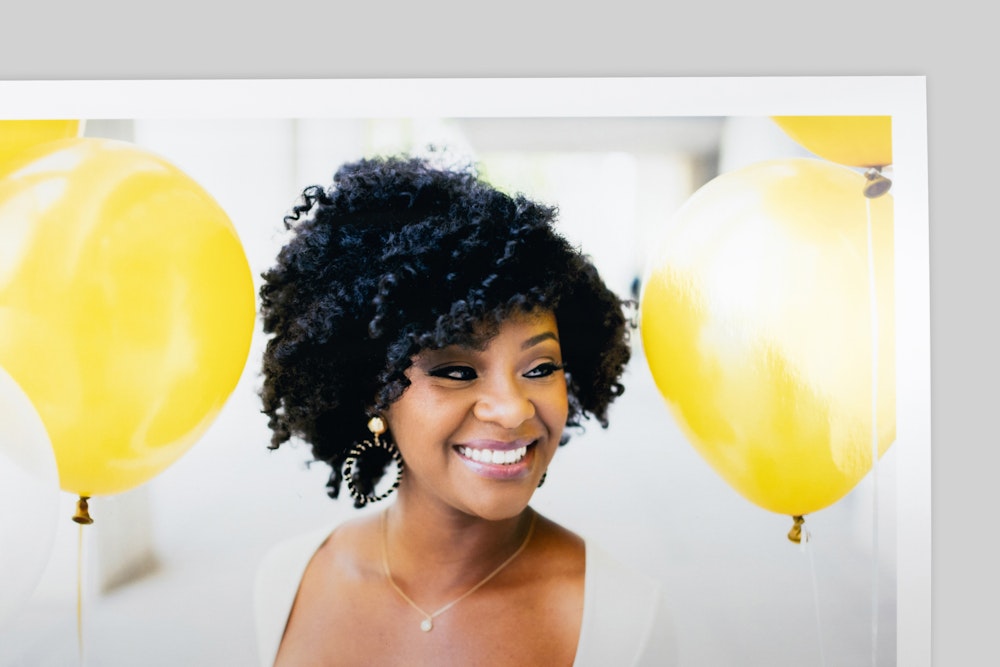 Corner of Pearl Photo Print with border, featuring a portrait of a smiling woman with curly hair amid bright yellow balloons. Strong reflection and glossy finish visible on paper surface.