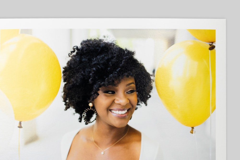 Corner of Photo Rag Baryta Fine Art Print with border, featuring portrait of a smiling woman with curly hair amid bright yellow balloons. Slight sheen visible on paper surface.