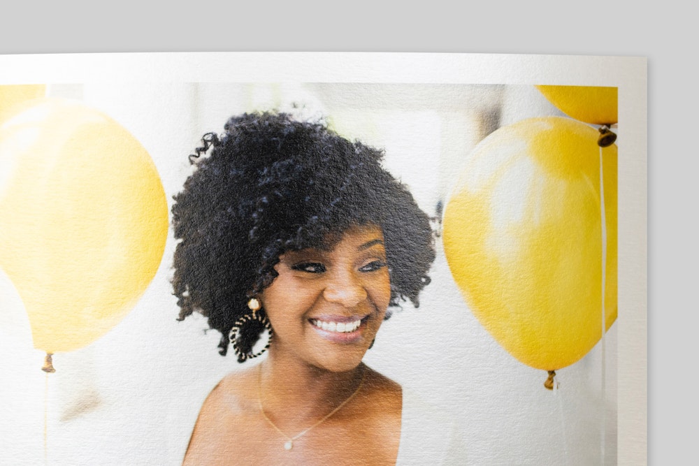 Corner of Photo Rag Metallic Fine Art Print with border, featuring portrait of a smiling woman with curly hair amid bright yellow balloons. Shimmery finish visible on paper surface.