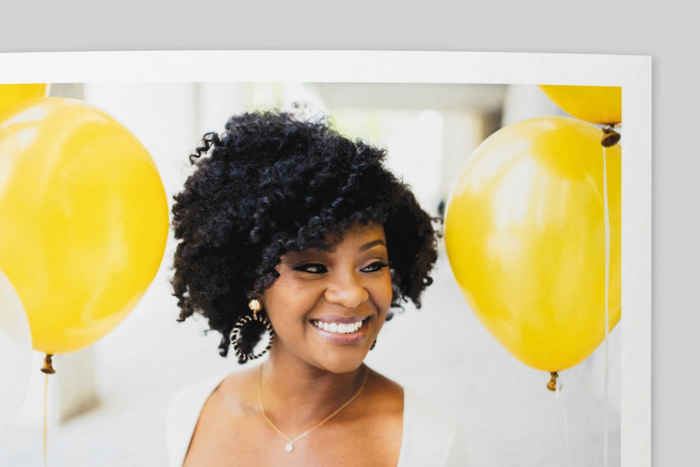 Corner of Torchon Fine Art Print with border, featuring a portrait of a smiling woman with curly hair amid bright yellow balloons. Coarse texture visible on paper surface.