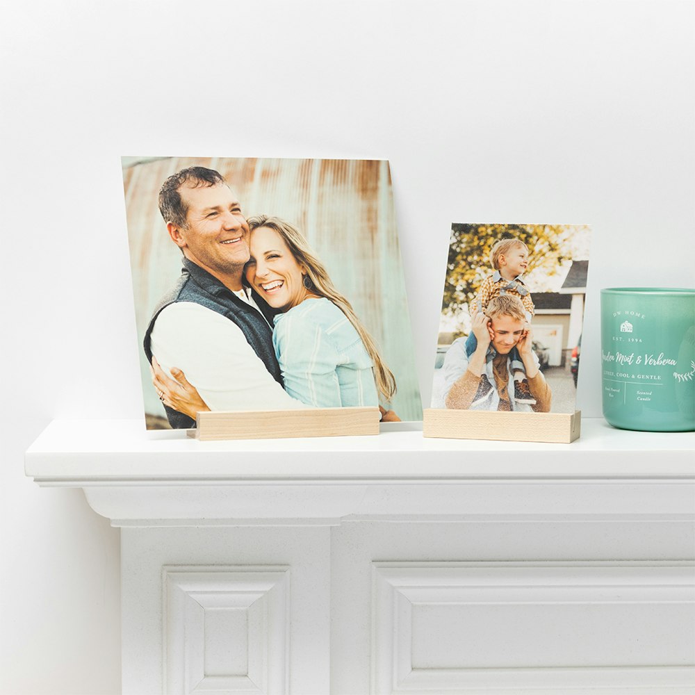 Two small photographic prints on light wooden stands, displaying a smiling couple and a playful family moment, sit on a white mantelpiece alongside a teal candle, in a softly lit, cozy setting.