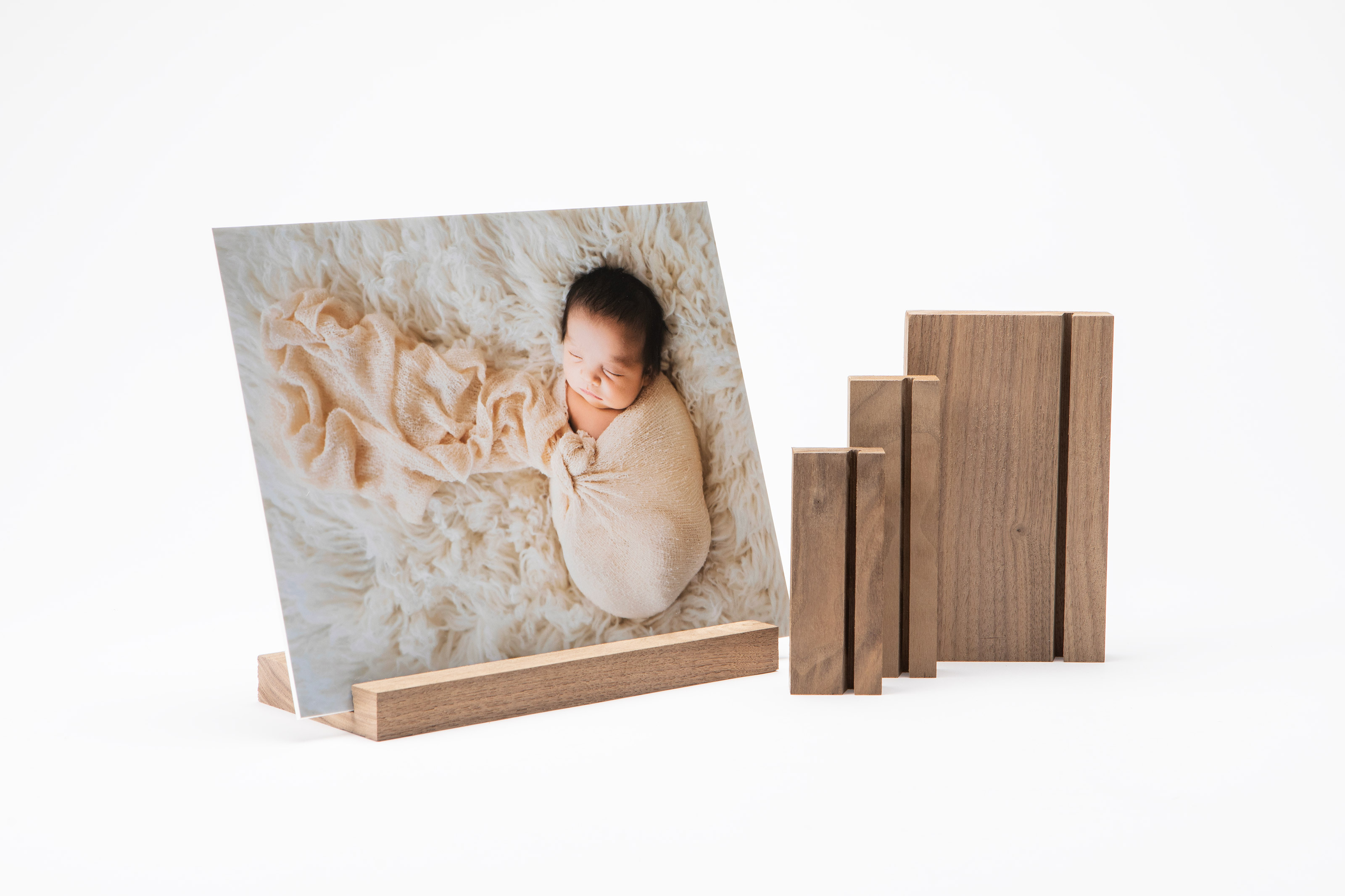 A mounted photo print of a sleeping baby on a walnut Wood Display Stand next to a set of three stand size options on a plain white background.